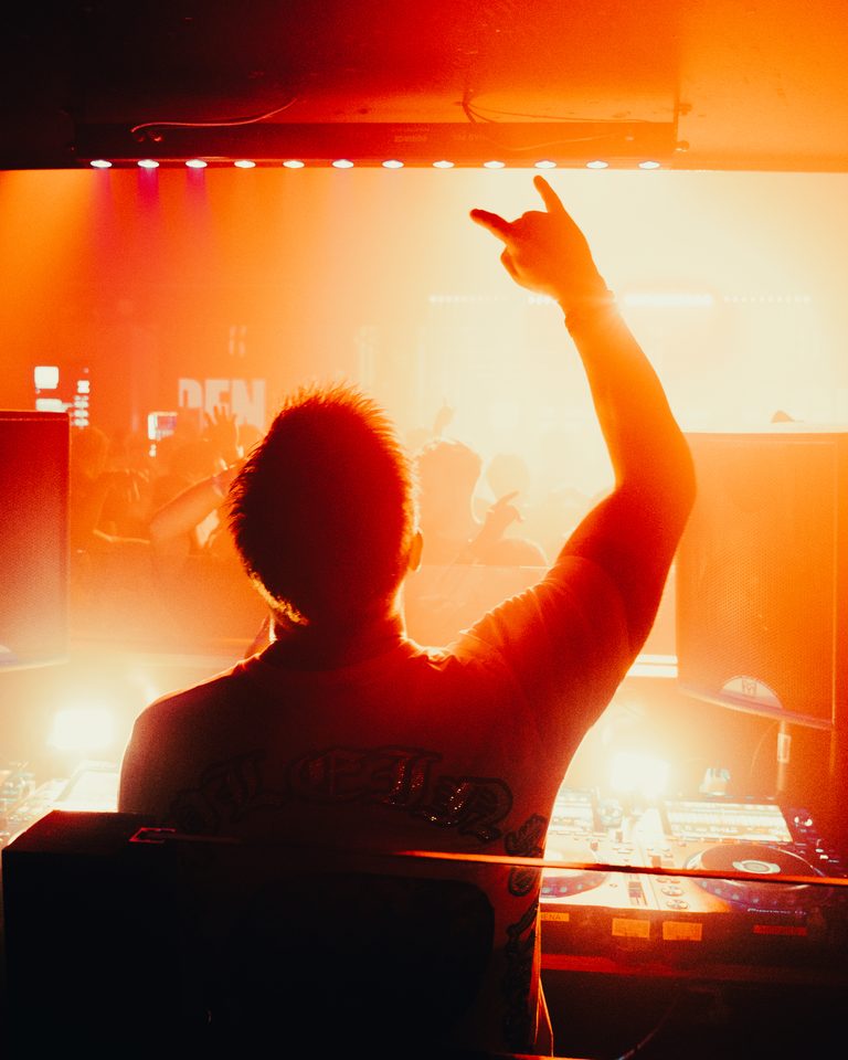 Silhouette of a DJ raising a hand, illuminated by stage lights.