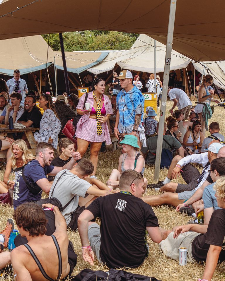 People gathered under tents on dry grass, some seated, some standing, in bright sun.