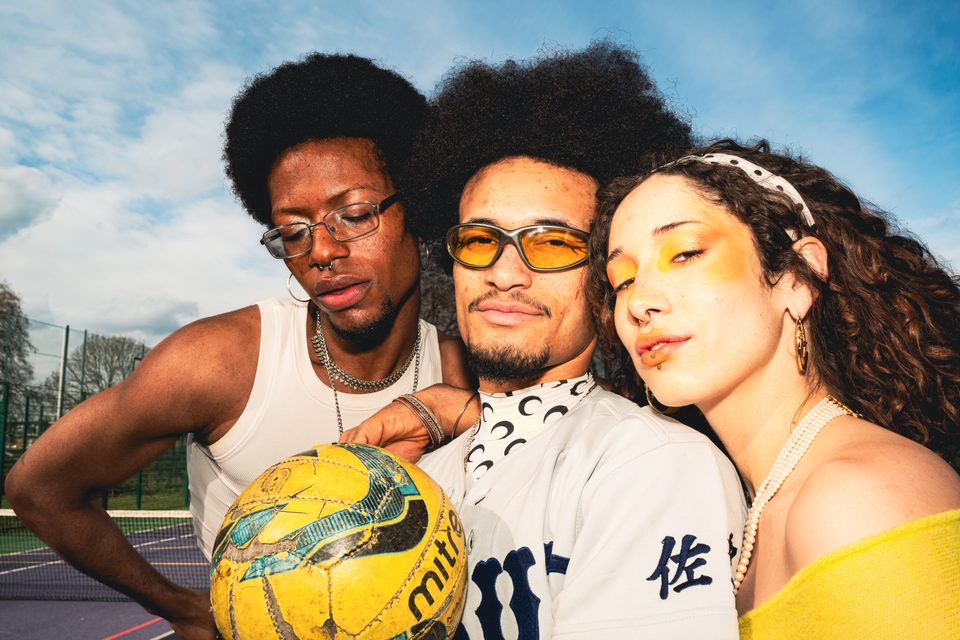 Three people posing with a worn soccer ball under bright sky.